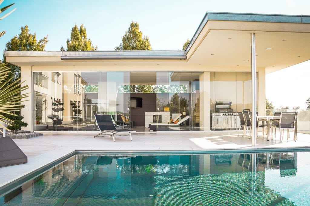 Off-white exterior of mid-century modern home with large siding glass doors, patio furniture and pool.