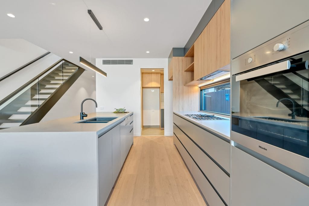 Modern minimalist kitchen with light grey cupboards, light wood upper cupboards, built-in oven and large island with sink.